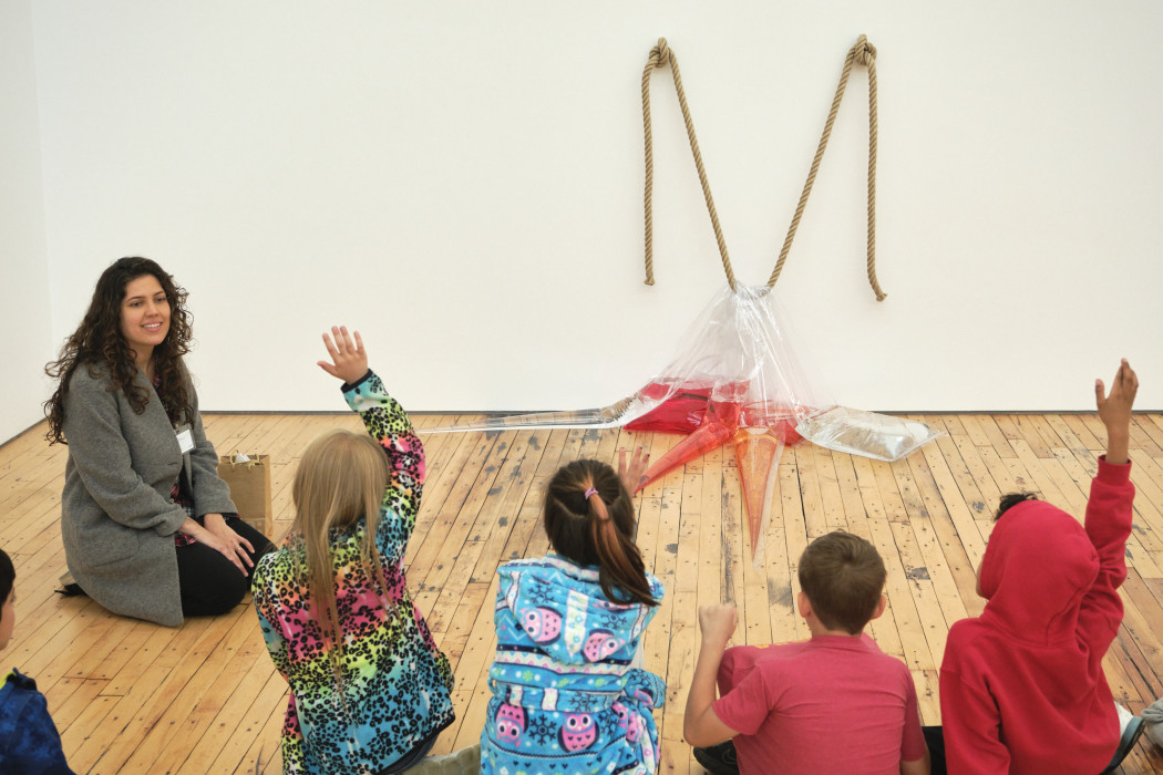 A group of children sit in front of an artwork made from plastic sheets and color water. A Dia educator sits between them and the work. One child has their hand raised to ask a question. 